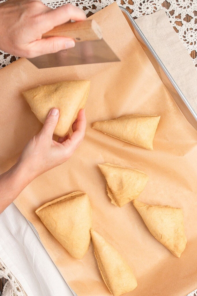 Portioned triangles of hot dog bun dough made with freshly milled flour on a parchment-lined baking sheet with a bench scraper shaping one piece