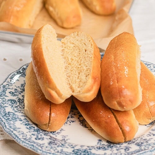 A split hot dog bun made with freshly milled flour showing its soft fluffy crumb, stacked with more buns on a blue and white floral plate