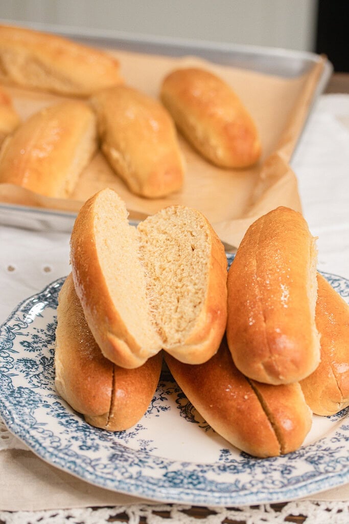 A split hot dog bun made with freshly milled flour showing its soft fluffy crumb, stacked with more buns on a blue and white floral plate