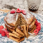 Stack of pancakes made with freshly milled flour on a blue and white floral plate, cut into a wedge to show the fluffy interior, topped with powdered sugar and fresh raspberries, with a glass of maple syrup in the background.