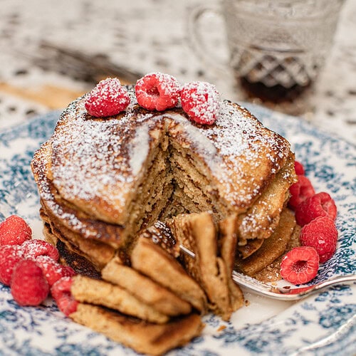 Stack of pancakes made with freshly milled flour on a blue and white floral plate, cut into a wedge to show the fluffy interior, topped with powdered sugar and fresh raspberries, with a glass of maple syrup in the background.