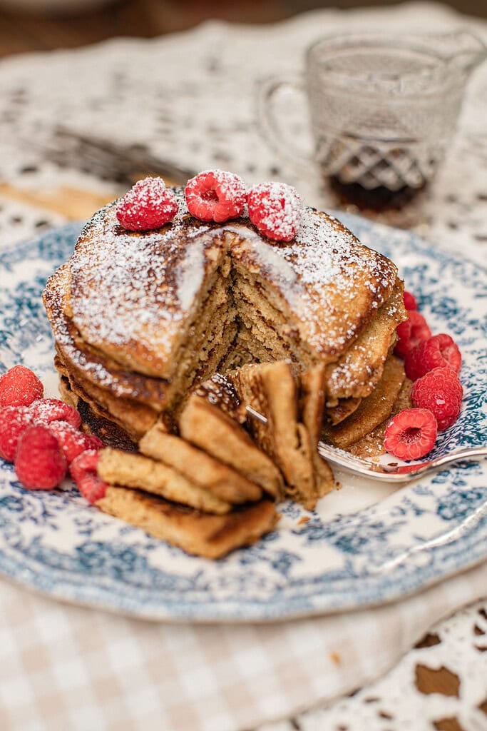 Stack of pancakes made with freshly milled flour on a blue and white floral plate, cut into a wedge to show the fluffy interior, topped with powdered sugar and fresh raspberries, with a glass of maple syrup in the background.