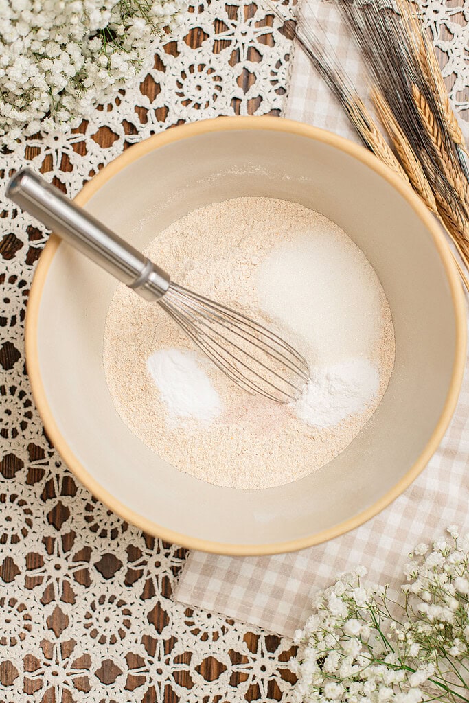 Overhead view of freshly milled flour, sugar, baking powder, and baking soda whisked together in a beige mixing bowl, with dried wheat stalks resting alongside on a checked cloth.