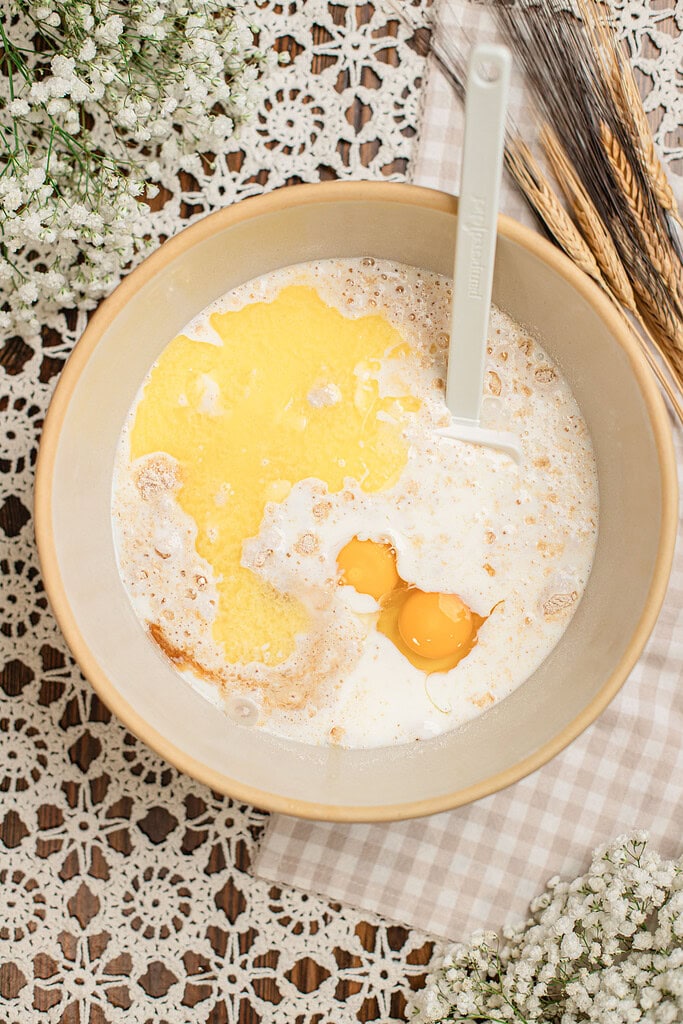 Overhead view of melted butter, milk, and eggs added to freshly milled flour in a mixing bowl, not yet stirred together, with a spatula resting in the bowl and dried wheat stalks nearby.