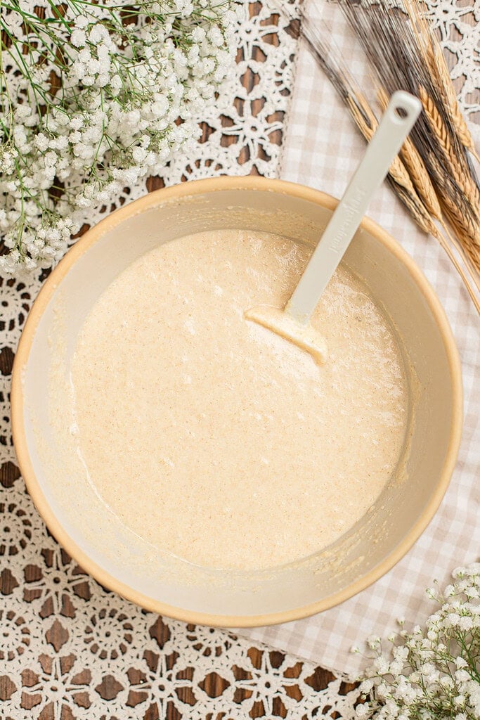 Overhead view of smooth pancake batter made with freshly milled flour, fully mixed in a beige bowl with a spatula resting inside, surrounded by baby's breath and dried wheat stalks.