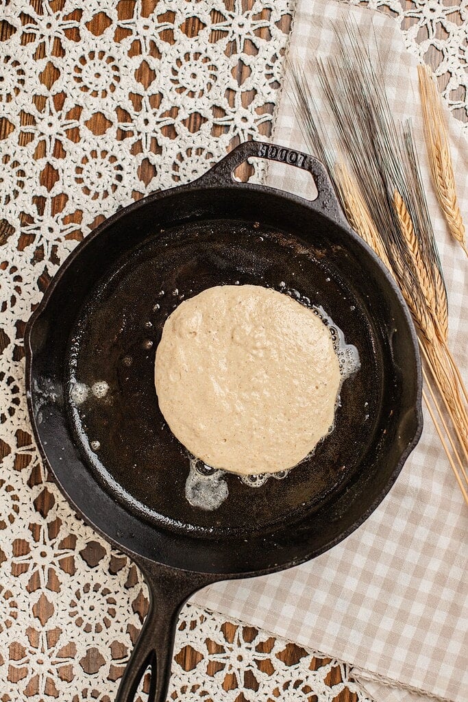 Overhead view of pancake batter poured into a hot cast iron skillet, not yet showing bubbles, with dried wheat stalks resting beside the pan.