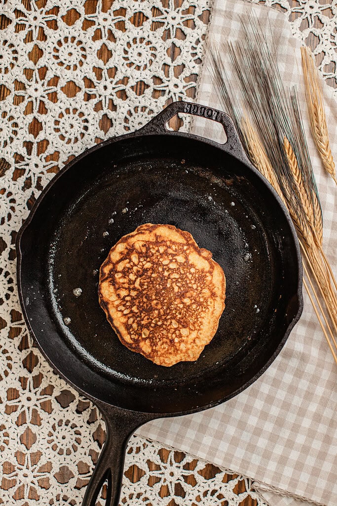 Overhead view of a golden brown pancake made with freshly milled flour cooking in a cast iron skillet, showing a bubbled surface ready to flip.