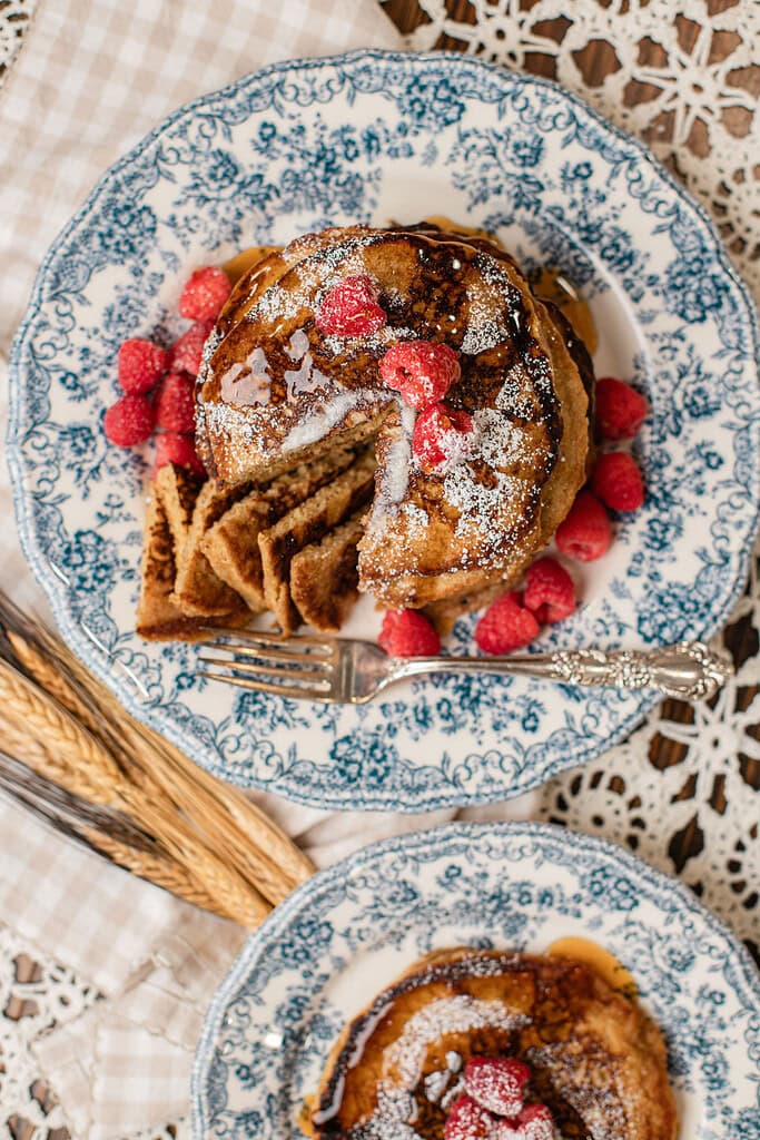 Overhead view of a stack of freshly milled flour pancakes cut into wedges on a blue and white floral plate, drizzled with maple syrup, dusted with powdered sugar, and topped with fresh raspberries, with a second plate of pancakes and dried wheat stalks nearby.