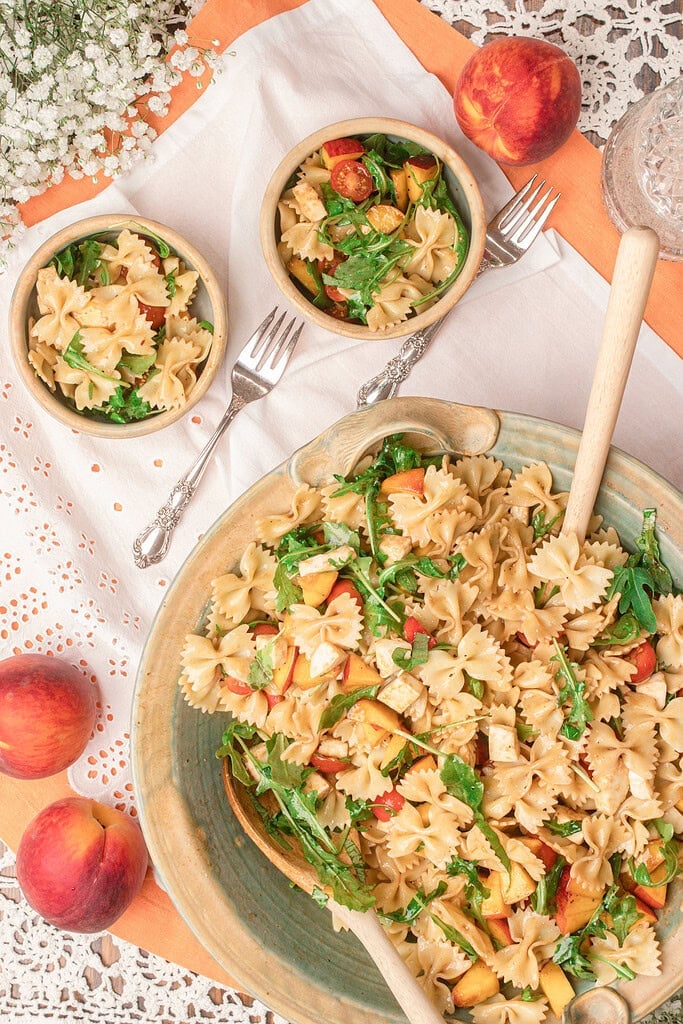 Overhead view of peach caprese pasta salad in a large serving bowl with individual bowls and fresh peaches