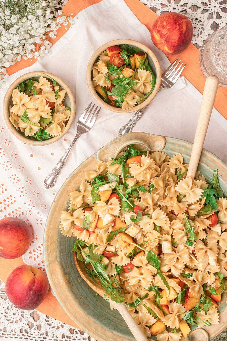 Overhead view of peach caprese pasta salad in a large serving bowl with individual bowls and fresh peaches