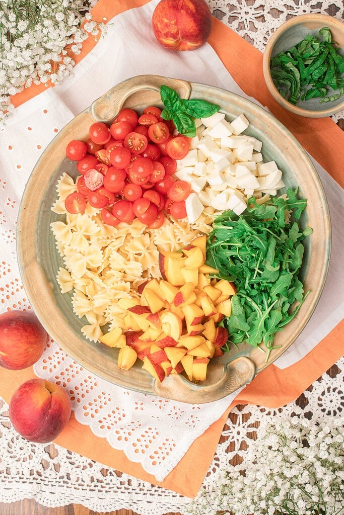 Bowl of peach caprese pasta salad ingredients arranged by section before mixing, with cherry tomatoes, mozzarella, arugula, and peaches
