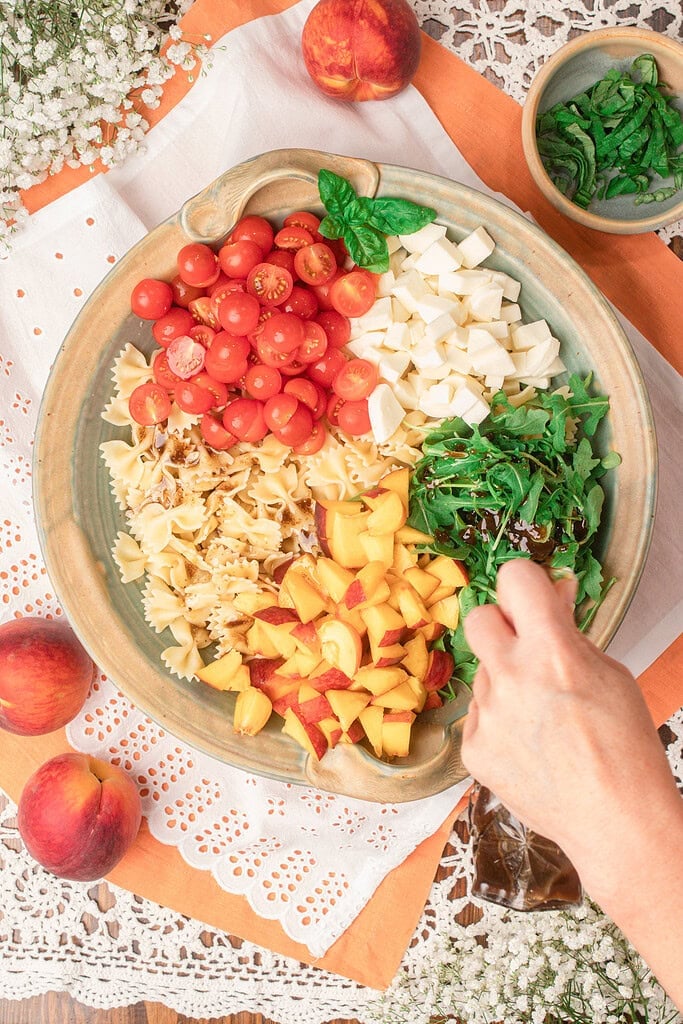 Drizzling balsamic vinaigrette over peach caprese pasta salad ingredients arranged in a serving bowl