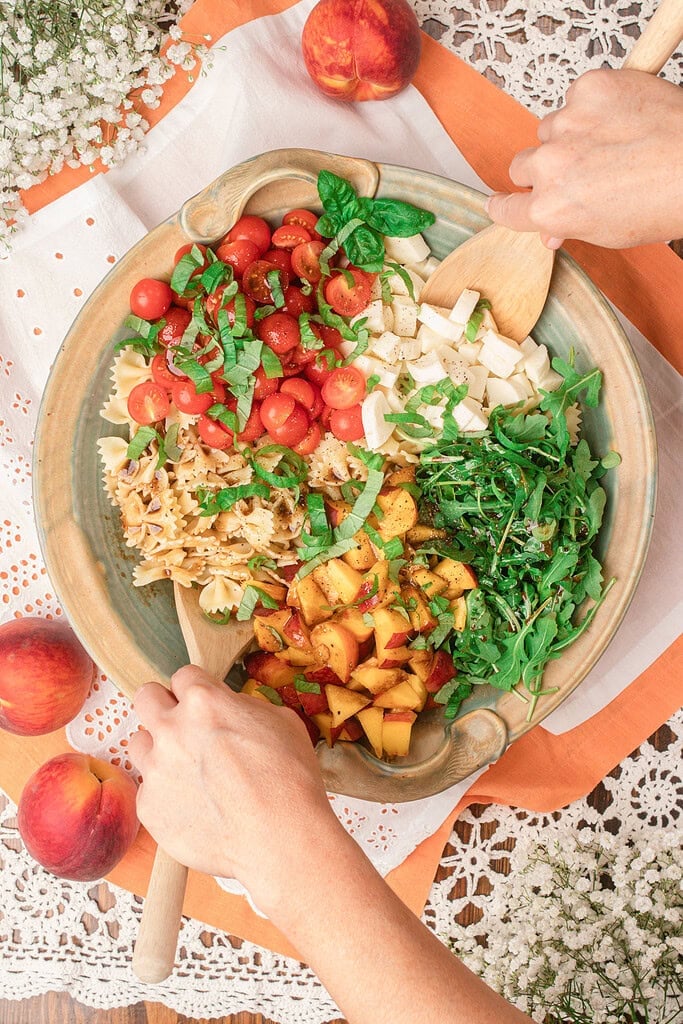 Tossing peach caprese pasta salad with a wooden spoon in a large ceramic serving bowl