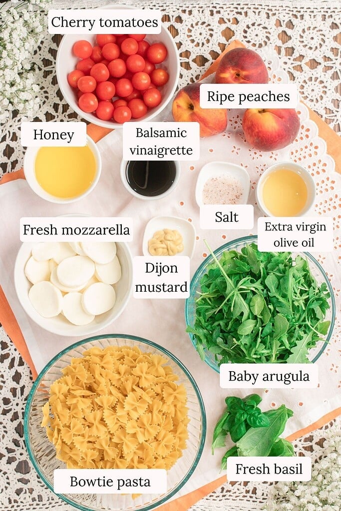 Overhead ingredient photo for peach Caprese pasta salad featuring bowtie pasta, ripe peaches, cherry tomatoes, fresh mozzarella, baby arugula, basil, Dijon mustard, balsamic vinaigrette, olive oil, honey, and salt.