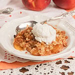 Homemade peach crisp with fresh milled flour topped with a scoop of vanilla ice cream in a white dessert bowl with fresh peaches in the background.