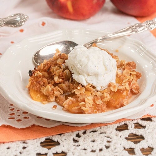 Homemade peach crisp with fresh milled flour topped with a scoop of vanilla ice cream in a white dessert bowl with fresh peaches in the background.