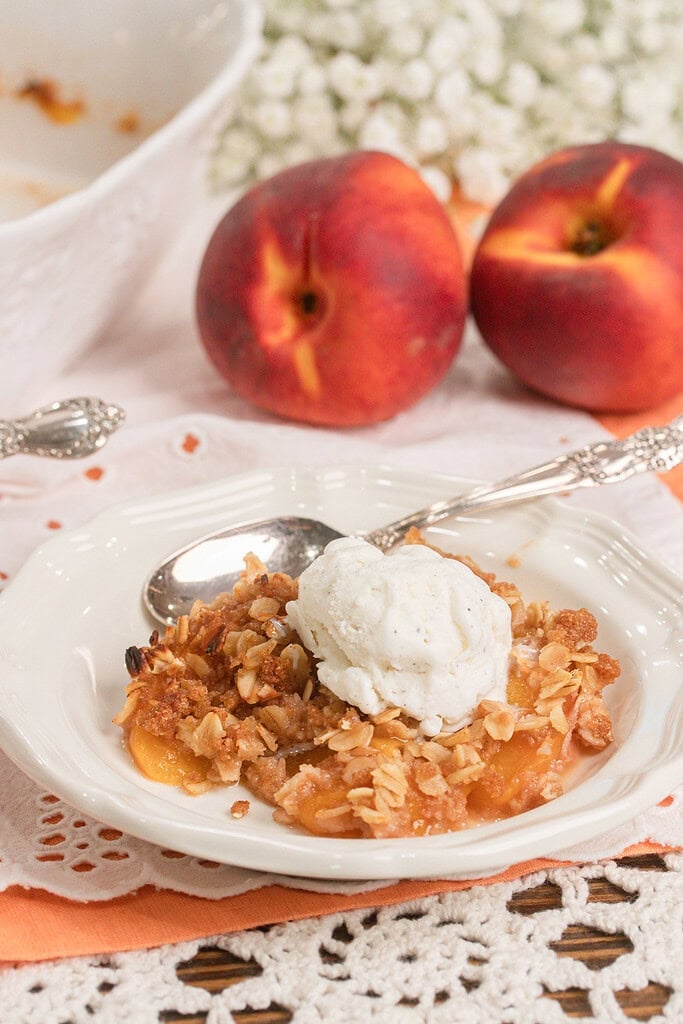 Homemade peach crisp with fresh milled flour topped with a scoop of vanilla ice cream in a white dessert bowl with fresh peaches in the background.