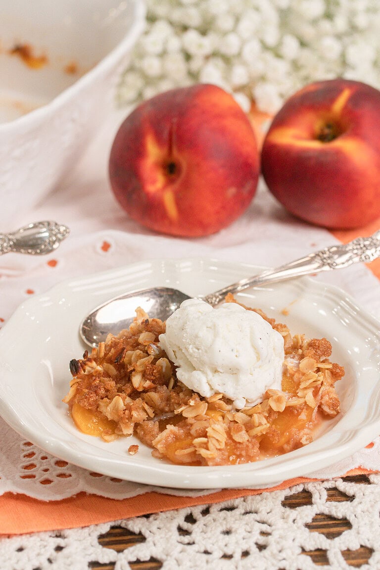 Homemade peach crisp with fresh milled flour topped with a scoop of vanilla ice cream in a white dessert bowl with fresh peaches in the background.