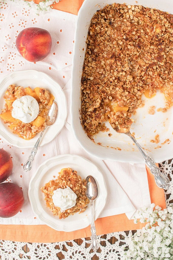 Overhead view of homemade peach crisp with fresh milled flour served with vanilla ice cream beside a baking dish of peach crisp and fresh peaches.