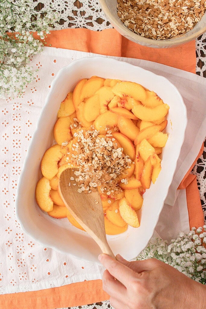 Sprinkling fresh milled flour oat crisp topping over sliced peaches in a baking dish before baking homemade peach crisp.