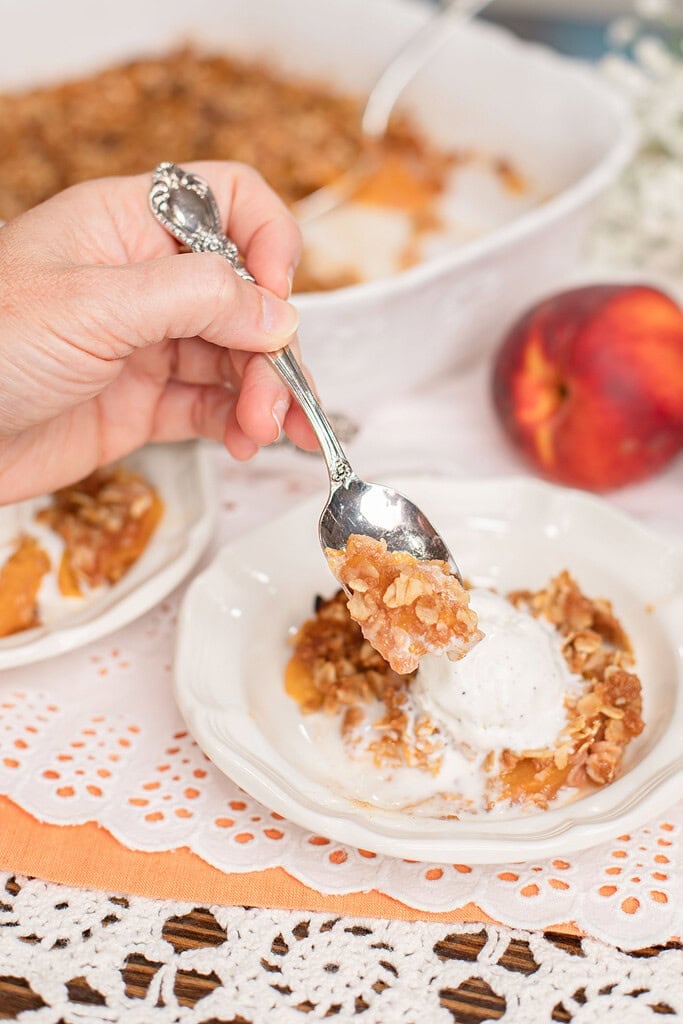 Spoonful of homemade peach crisp with fresh milled flour oat topping and vanilla ice cream lifted from a white dessert bowl.