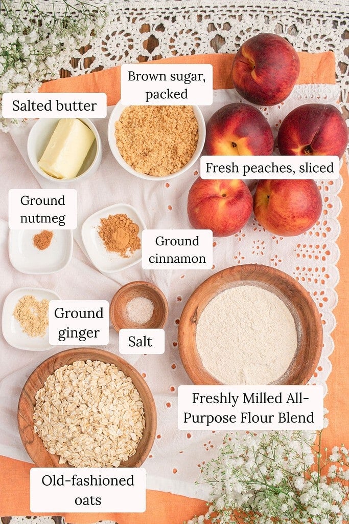 Flat lay of ingredients for peach crisp with fresh milled flour including sliced fresh peaches, fresh milled all-purpose flour blend, old-fashioned oats, brown sugar, butter, cinnamon, ginger, nutmeg, and salt on a cozy cottage-style backdrop.