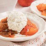 Homemade peach crumble made with fresh milled flour topped with a scoop of vanilla ice cream in a white dessert bowl with fresh peaches in the background.
