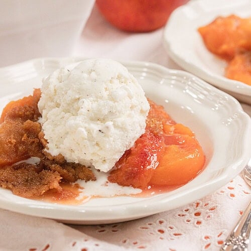 Homemade peach crumble made with fresh milled flour topped with a scoop of vanilla ice cream in a white dessert bowl with fresh peaches in the background.