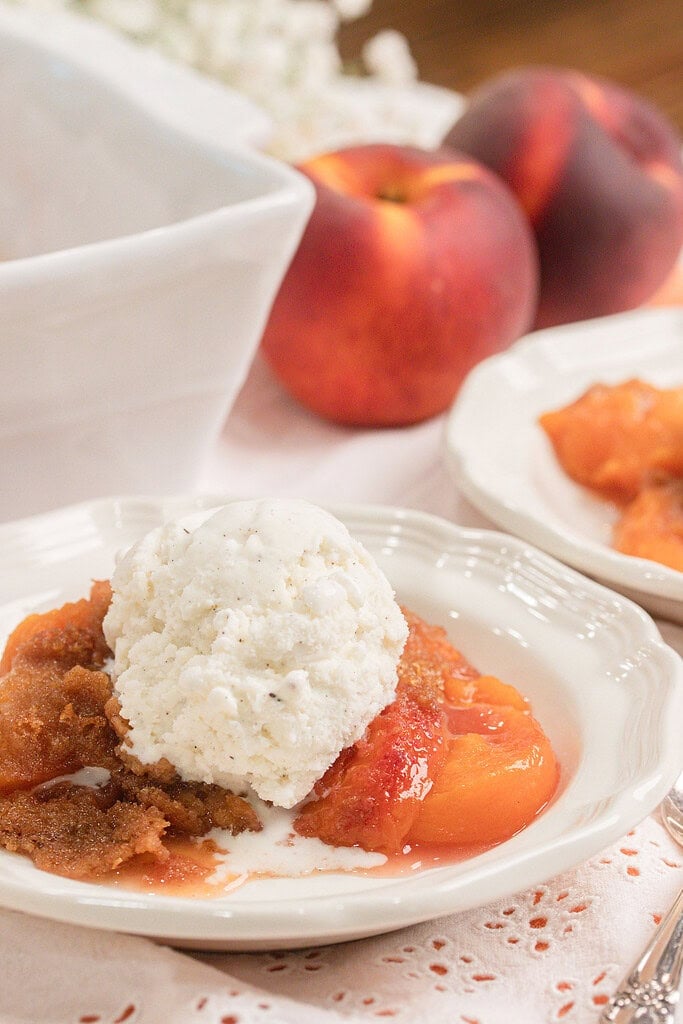 Homemade peach crumble made with fresh milled flour topped with a scoop of vanilla ice cream in a white dessert bowl with fresh peaches in the background.
