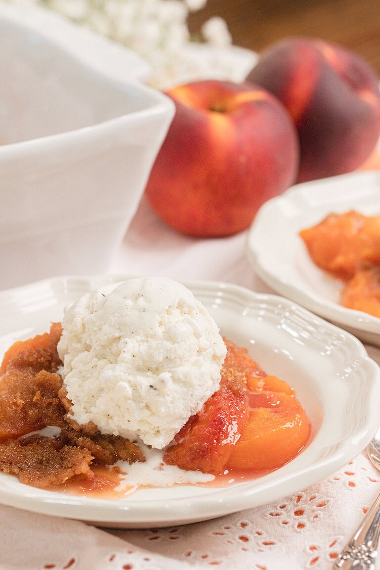 Homemade peach crumble made with fresh milled flour topped with a scoop of vanilla ice cream in a white dessert bowl with fresh peaches in the background.