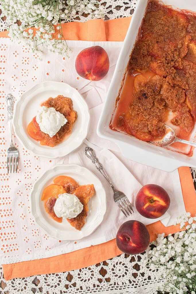 Overhead view of homemade peach crumble with fresh milled flour served with vanilla ice cream beside a baking dish of peach crumble and fresh peaches.
