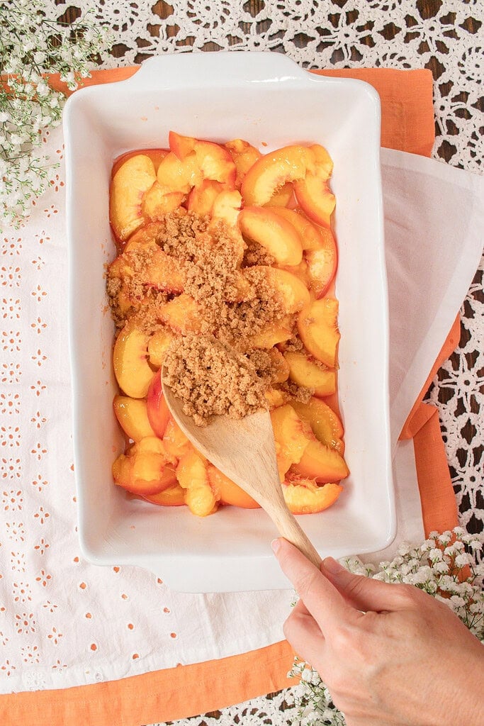Sprinkling fresh milled flour crumble topping over sliced peaches in a baking dish before baking homemade peach crumble.