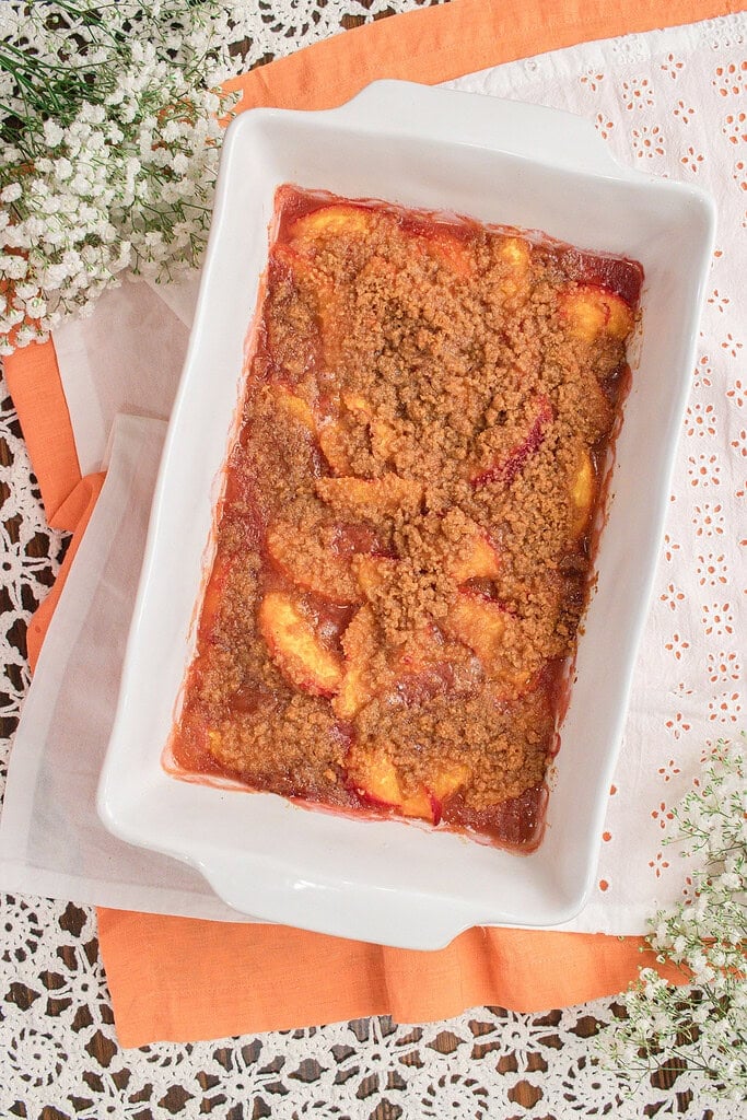 Fresh milled flour peach crumble baked until golden brown with bubbling peaches and a crisp cinnamon crumble topping in a white baking dish.