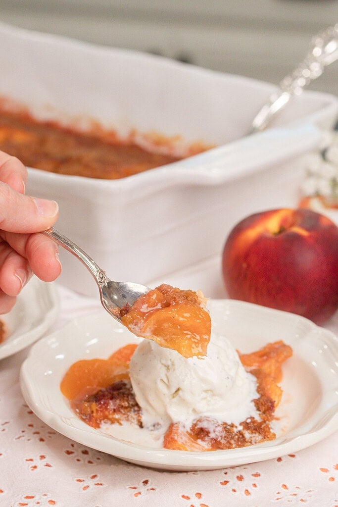 Spoonful of homemade peach crumble with fresh milled flour and vanilla ice cream lifted from a dessert bowl with baked peaches and cinnamon crumble topping.