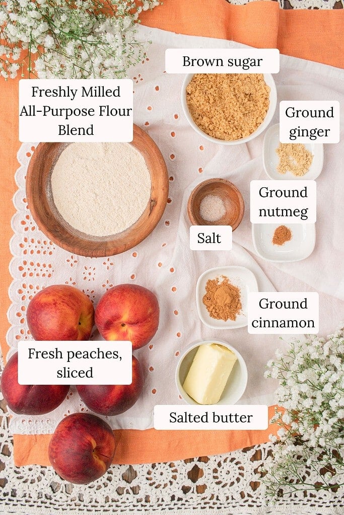 Ingredients for peach crumble with fresh milled flour arranged overhead with fresh peaches, fresh milled all-purpose flour blend, brown sugar, butter, cinnamon, ginger, nutmeg, and salt on a white linen cloth.