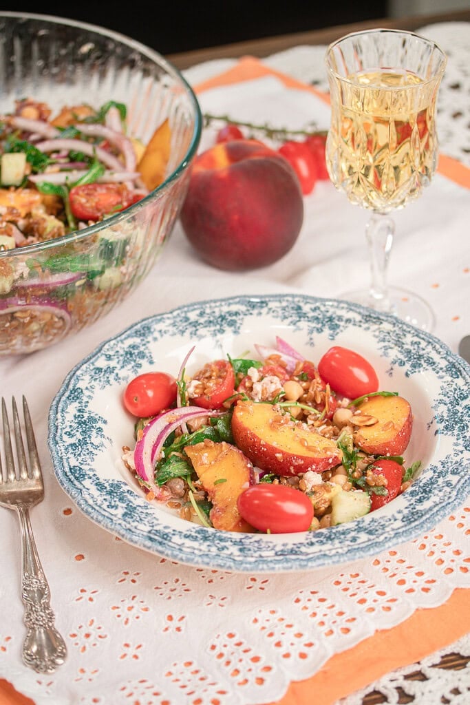 Bowl of homemade peach farro salad with fresh peaches, chickpeas, arugula, cherry tomatoes, red onion, feta, and toasted almonds served alongside a large salad bowl.
