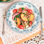 Overhead view of homemade peach farro salad served in a blue floral bowl with a large serving bowl, fresh peaches, cherry tomatoes, and a glass of white wine.