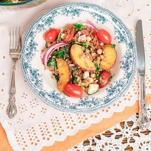 Overhead view of homemade peach farro salad served in a blue floral bowl with a large serving bowl, fresh peaches, cherry tomatoes, and a glass of white wine.