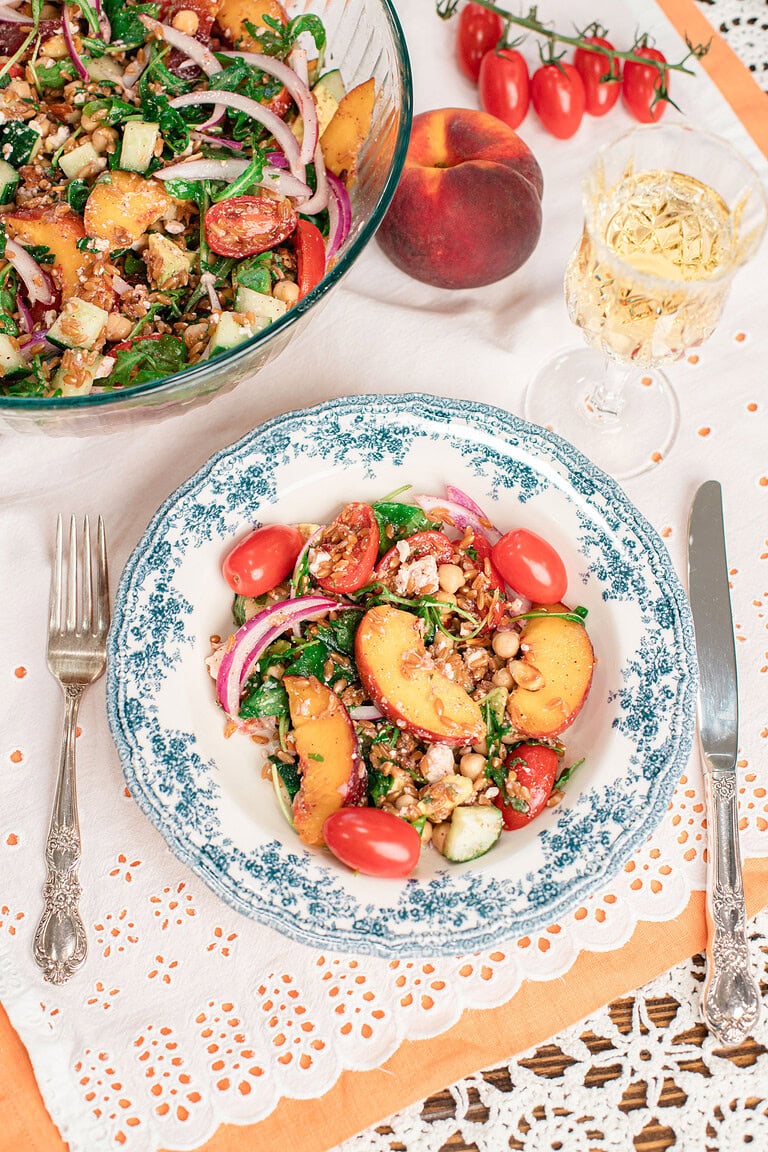 Overhead view of homemade peach farro salad served in a blue floral bowl with a large serving bowl, fresh peaches, cherry tomatoes, and a glass of white wine.