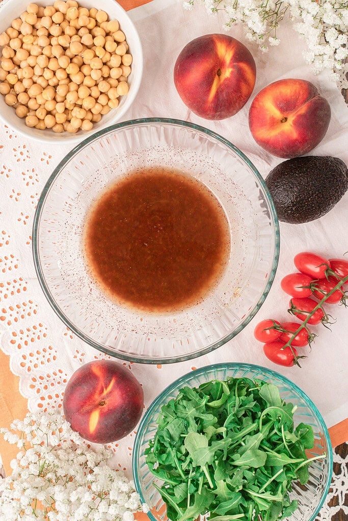 Homemade balsamic vinaigrette for peach farro salad in a glass mixing bowl surrounded by fresh peaches, arugula, chickpeas, avocado, and cherry tomatoes.