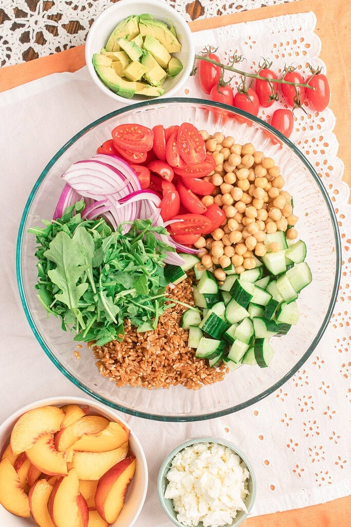 Fresh ingredients for homemade peach farro salad including cooked farro, arugula, chickpeas, cucumber, cherry tomatoes, red onion, avocado, peaches, and crumbled feta.