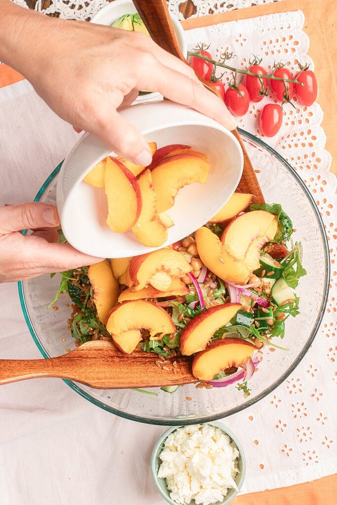 Adding fresh sliced peaches to a large bowl of homemade peach farro salad with farro, arugula, chickpeas, cucumber, tomatoes, and red onion.