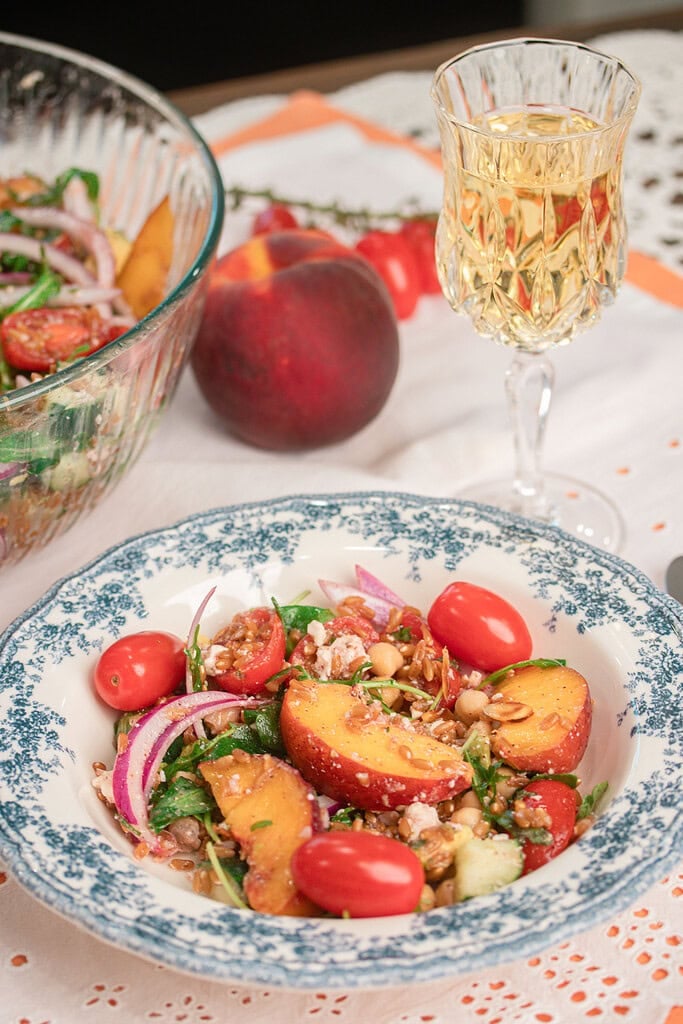 Overhead view of homemade peach farro salad served in a bowl with fresh peaches, chickpeas, arugula, cherry tomatoes, feta, toasted almonds, and red onion beside the serving bowl.