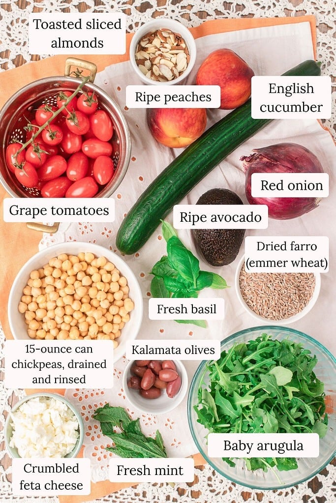 Ingredients for peach farro salad featuring ripe peaches, cooked farro, chickpeas, cucumber, grape tomatoes, avocado, feta cheese, Kalamata olives, arugula, basil, mint, toasted almonds, and red onion arranged in an overhead flat lay.