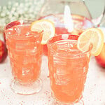 Two glasses of homemade peach lemonade served over ice with fresh lemon slices, with a pitcher of peach lemonade in the background.