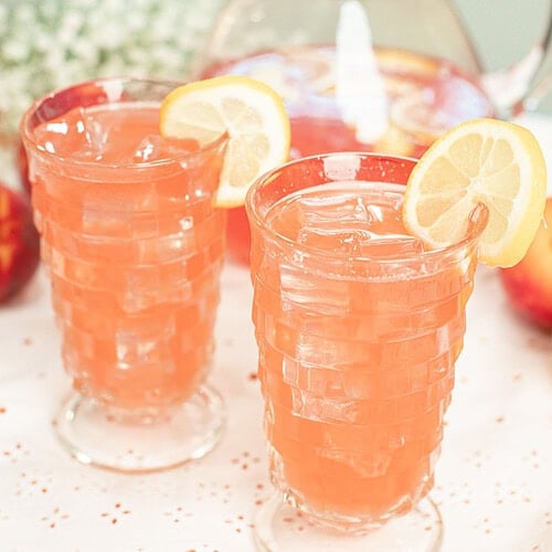 Two glasses of homemade peach lemonade served over ice with fresh lemon slices, with a pitcher of peach lemonade in the background.