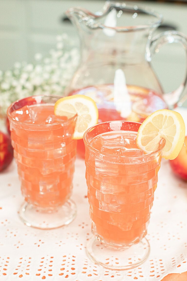 Two glasses of homemade peach lemonade served over ice with fresh lemon slices, with a pitcher of peach lemonade in the background.