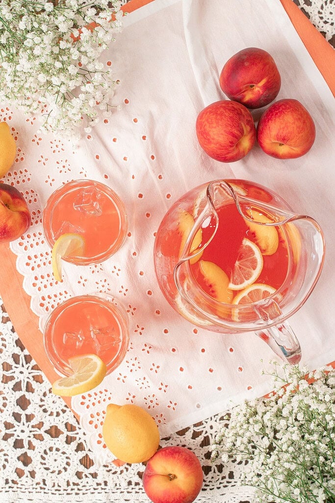 Overhead view of a pitcher of homemade peach lemonade with fresh peach slices and lemon slices, served alongside two glasses over ice.