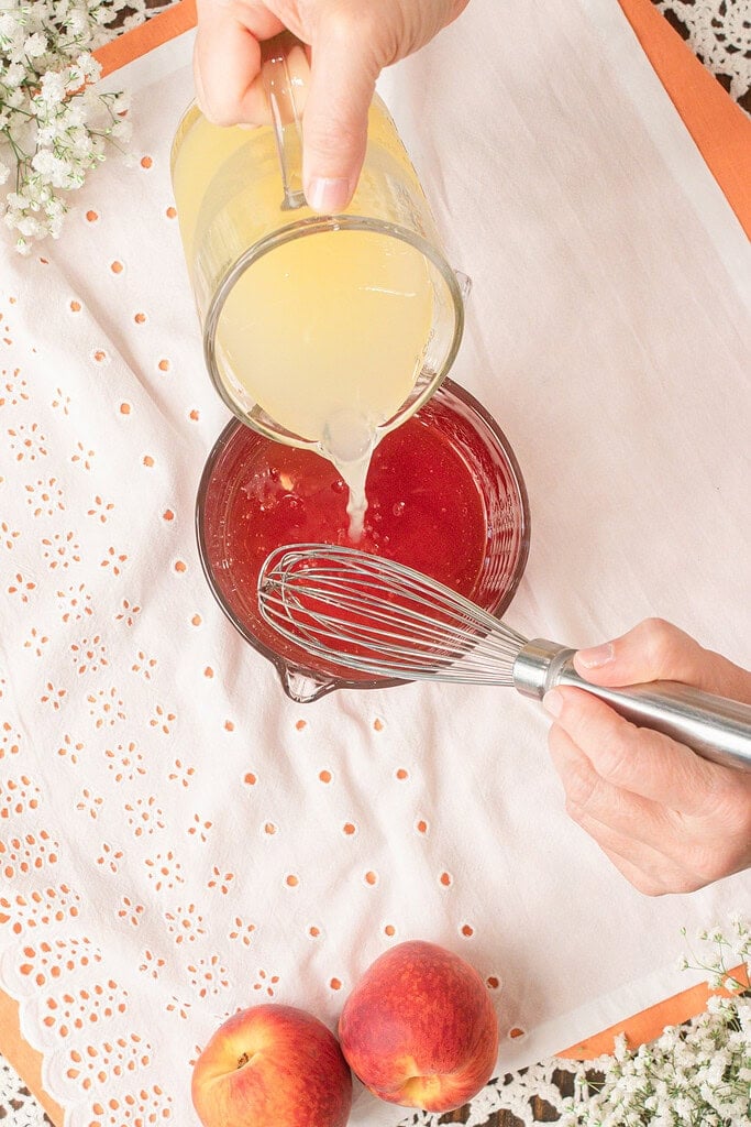 Pouring a homemade lemonade mixture into peach simple syrup while whisking to combine fresh peach lemonade ingredients.
