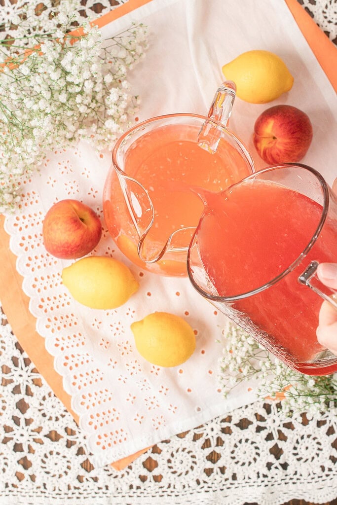 Pouring homemade peach lemonade into a glass pitcher for serving, surrounded by fresh peaches, lemons, and white flowers.
